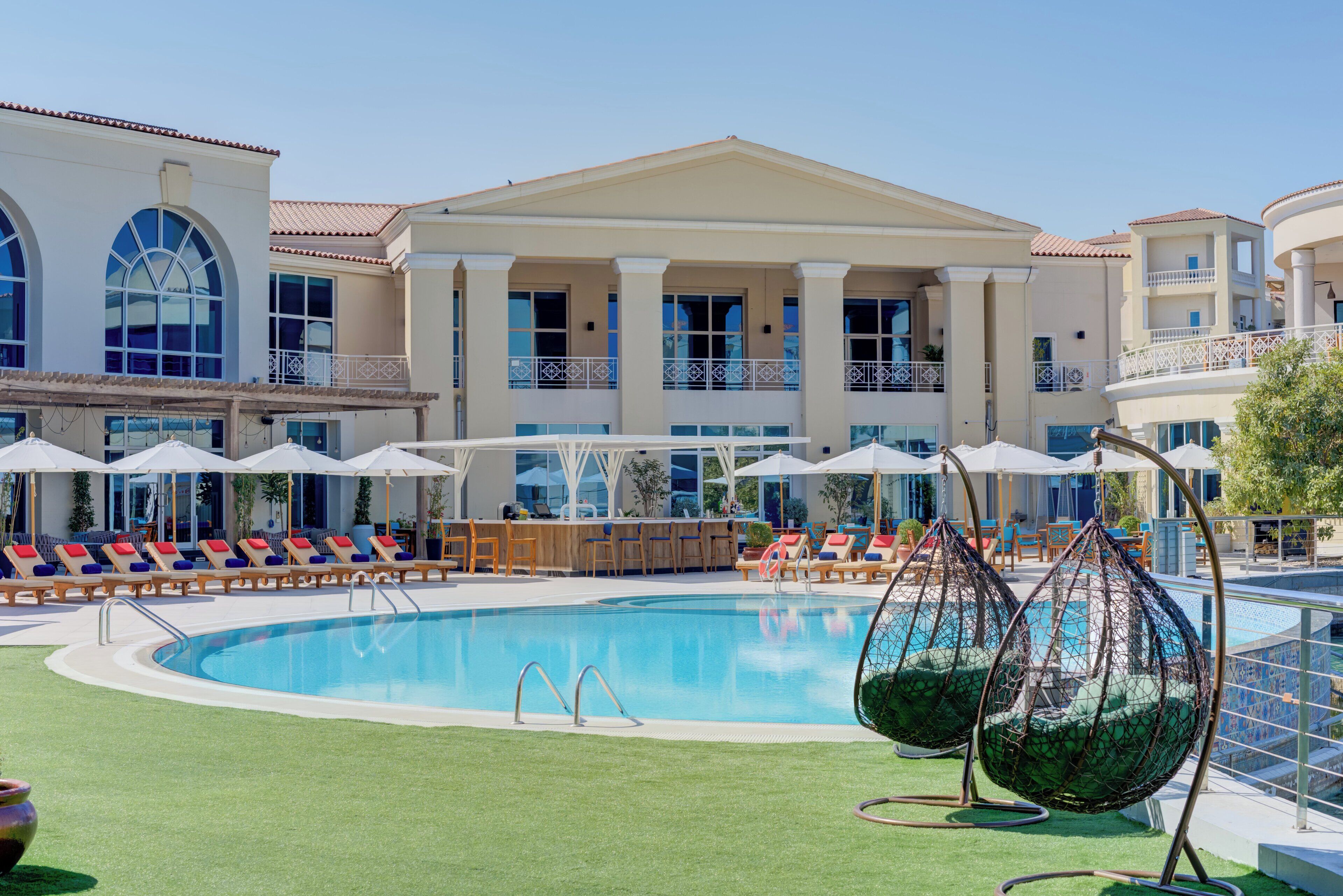 A pool area with lounge chairs, umbrellas, and a large building with columns.