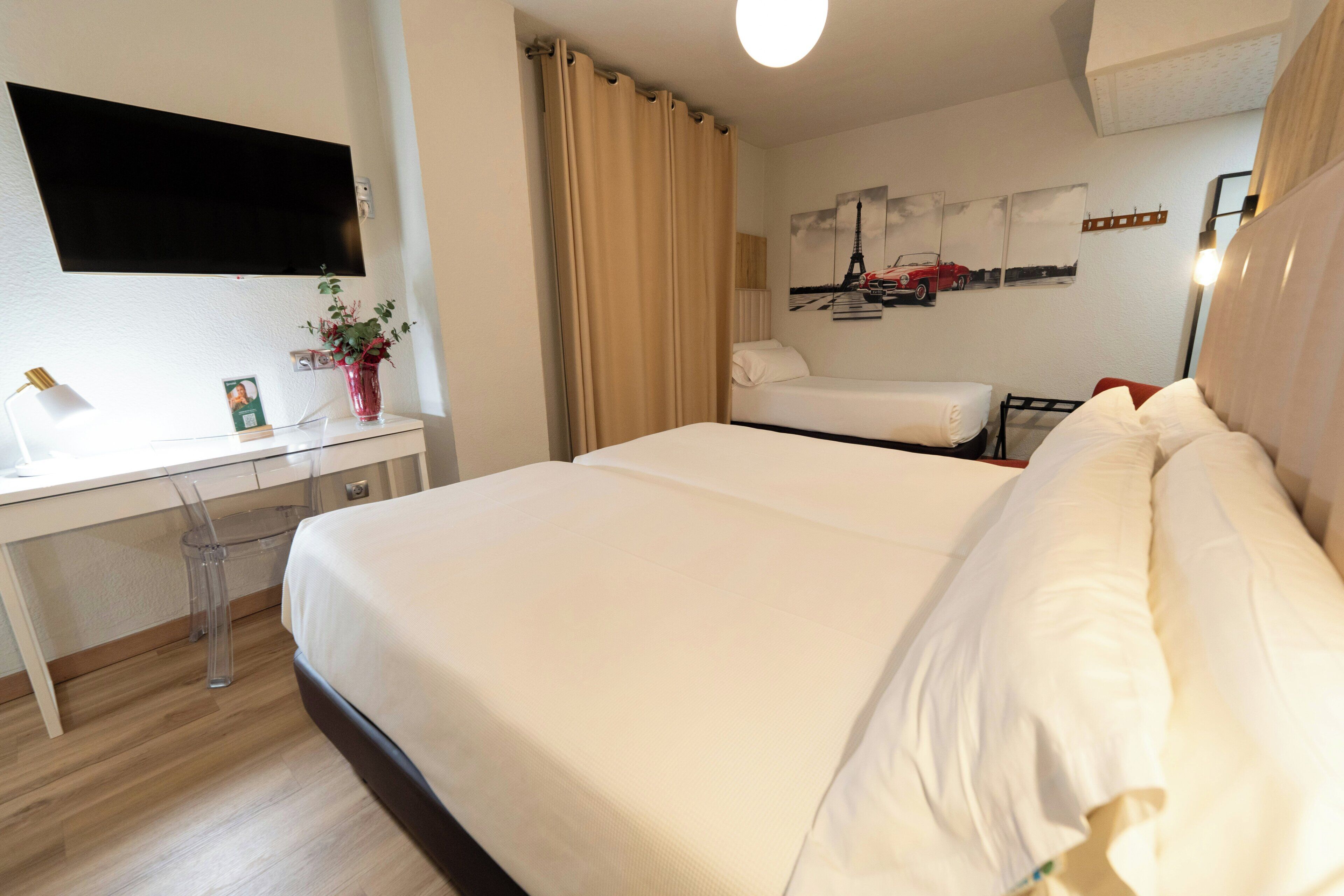 A hotel room with a bed, a desk, a chair, a TV mounted on the wall, and a window with curtains.
