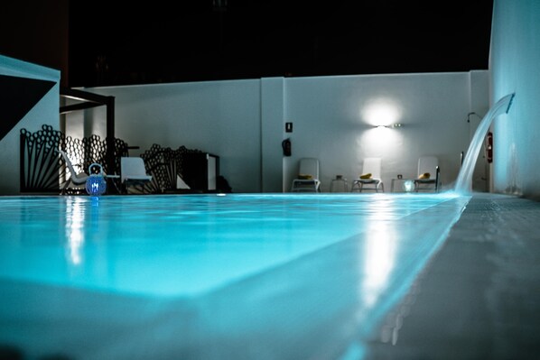 A swimming pool with a blue-lit edge, surrounded by white walls and a few chairs.
