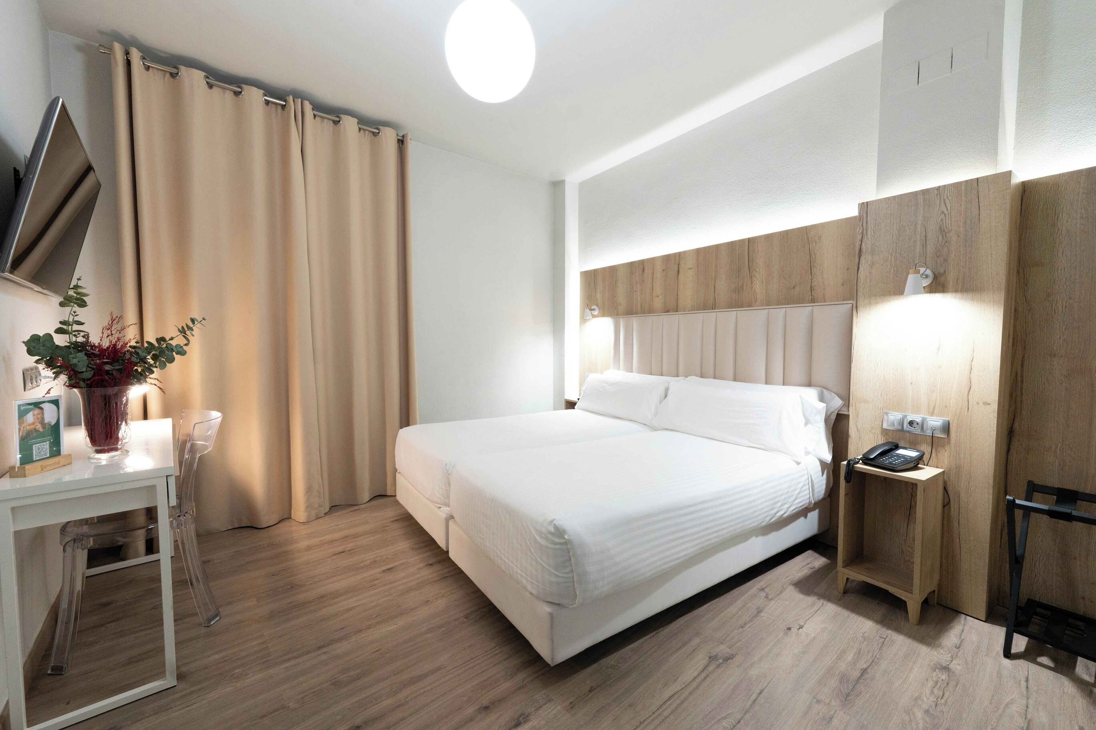 A hotel room with a bed, bedside table, lamp, and a potted plant.