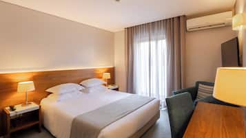 A hotel room with a large bed, a desk with a lamp, a chair, a TV, and an air conditioning unit.