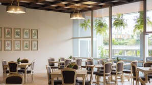Un comedor con varias mesas y sillas, ventanas amplias con plantas y cuadros en la pared.