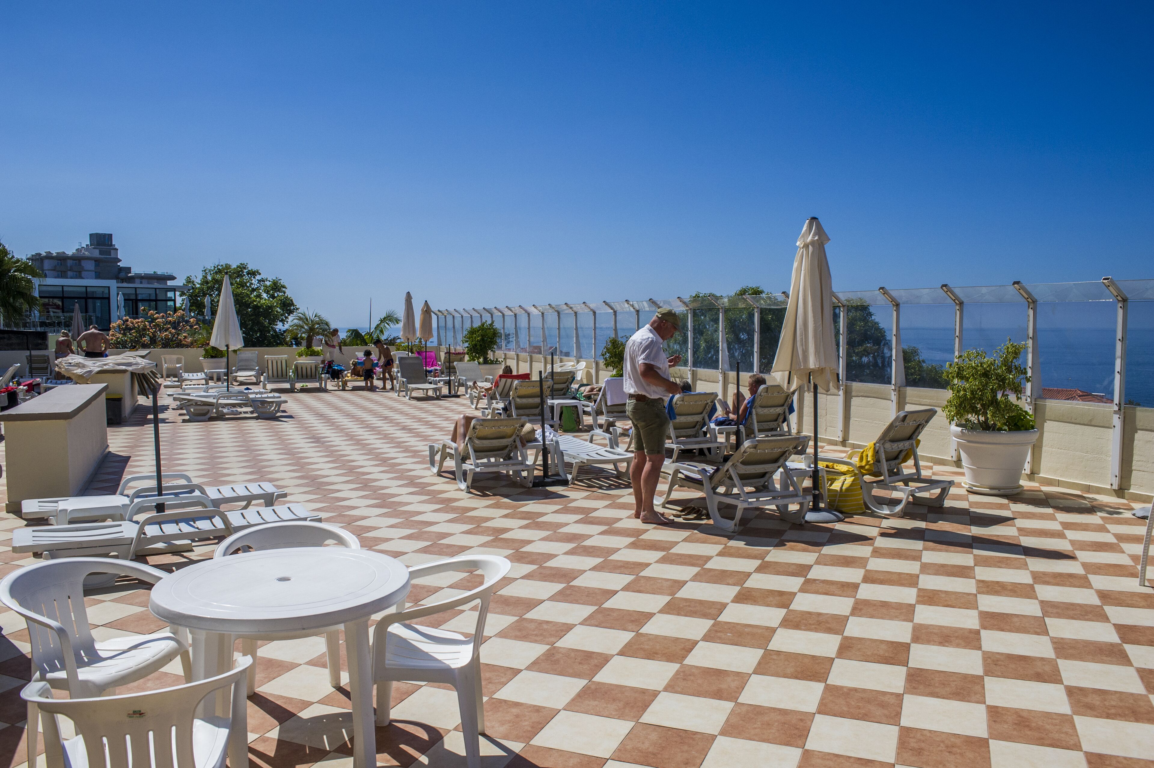Terraza al aire libre con piso a cuadros, sillones de descanso, mesas y sombrillas, con vista a una masa de agua.