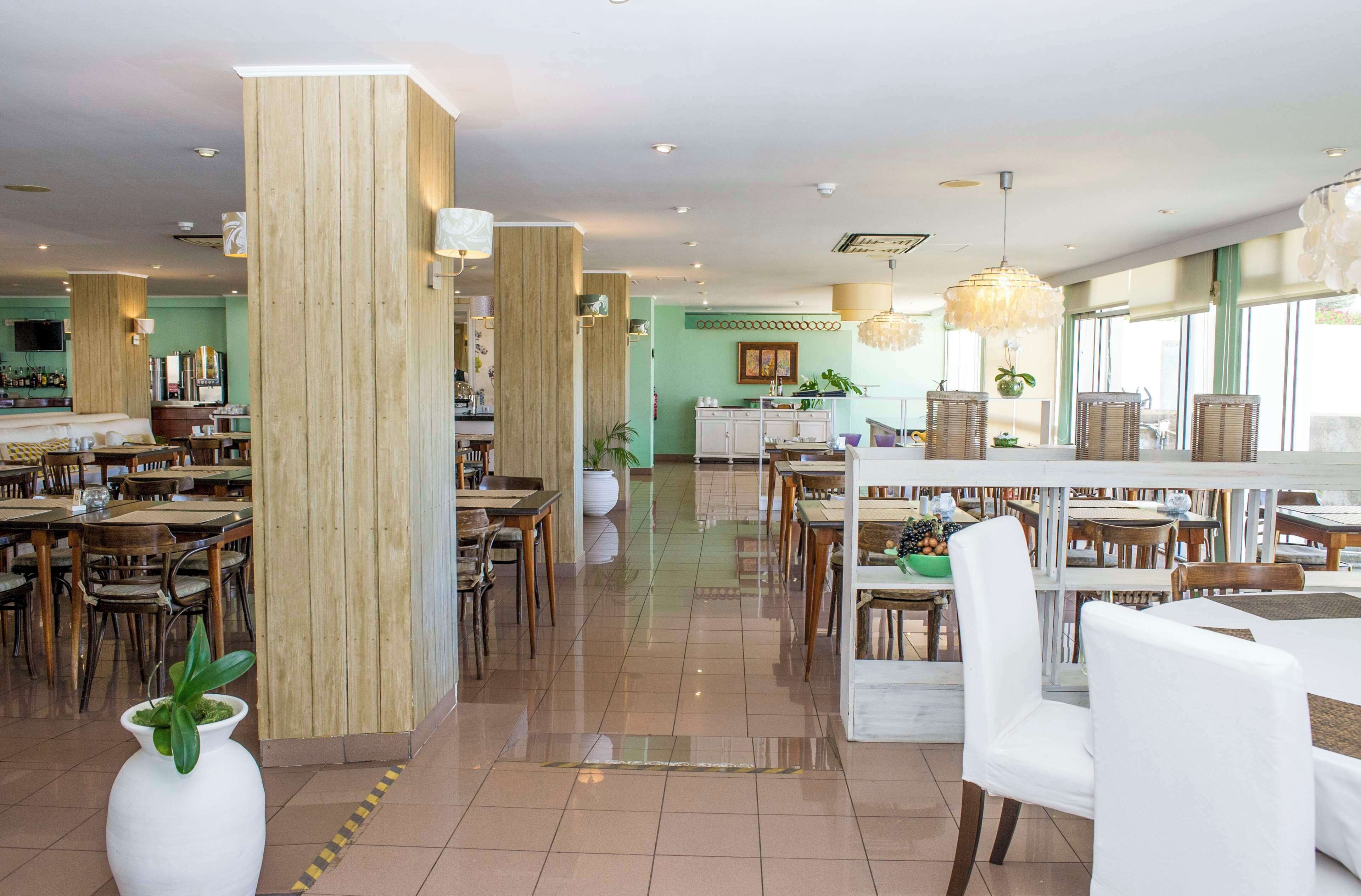 A spacious hotel dining area with multiple tables and chairs, wooden accents, and large windows.