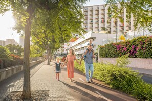 A family walking on a paved path with greenery and a building in the background. - Florasol Residence Hotel (Funchal)