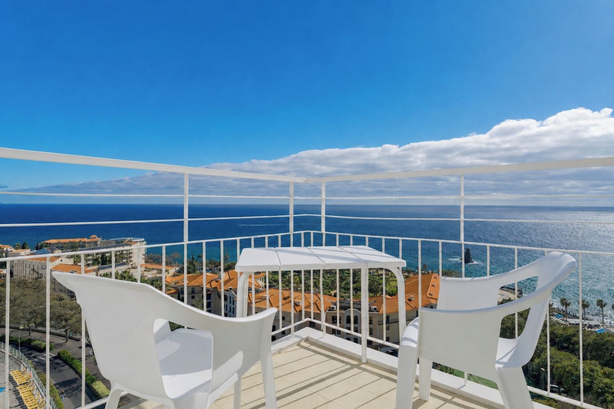 A balcony with a view of the sea, white chairs, and a table.