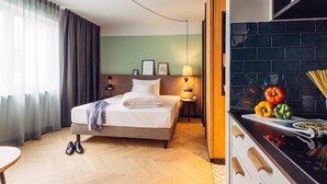 A hotel room with a bed, a nightstand, a lamp, a window with curtains, and a kitchen area with a tiled backsplash and a magazine on the counter. - Harry's Home Berlin Moabit (Berlin)