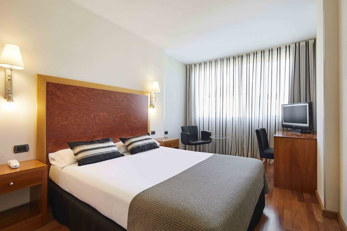 A hotel room with a large bed, two bedside lamps, a television, a desk, and a chair.