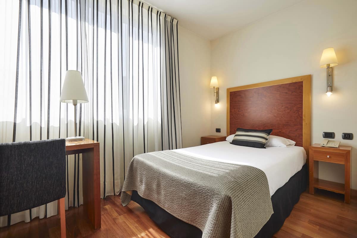 A hotel room with a bed, a desk with a lamp, a chair, and a nightstand with a phone.