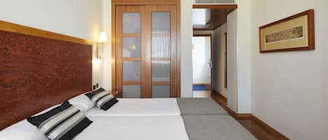 A hotel room with two beds, wooden headboard, a framed picture on the wall, and a hallway leading to another room.