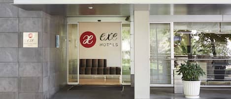 L'entrée de l'hôtel Exe Hotels Plaza Delicias se distingue par son design moderne, avec une porte en verre, un espace salon à l'intérieur et un pot de fleurs à l'extérieur.