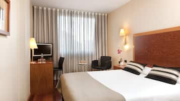 Habitación de hotel con una cama grande, un televisor, un escritorio con lámpara, una silla y una pequeña mesa con una flor.