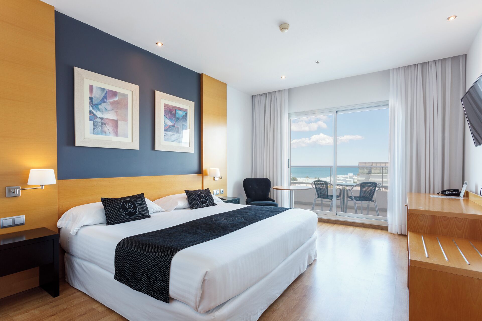 A hotel room with a large bed, two framed artworks, a desk with a chair, a small table with chairs, and a balcony with a view of the sea.