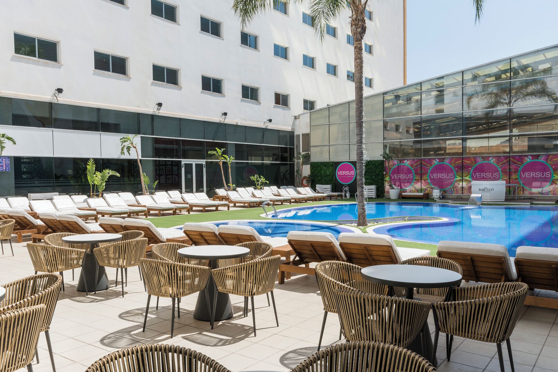 Zona de piscina de hotel con sillones y mesas, una alberca y un edificio con ventanas amplias y un letrero que dice 'VENSUS'.