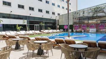 Zona de piscina de hotel con sillones y mesas, una alberca y un edificio con ventanas amplias y un letrero que dice 'VENSUS'.