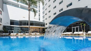 Una piscina d'hotel moderna amb cascada, gandules i un edifici d'altes plantes amb grans finestres.