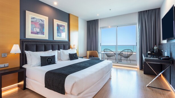 Habitació d'hotel moderna amb un llit gran, una taula de treball amb televisió de pantalla plana i un balcó amb vista al mar.