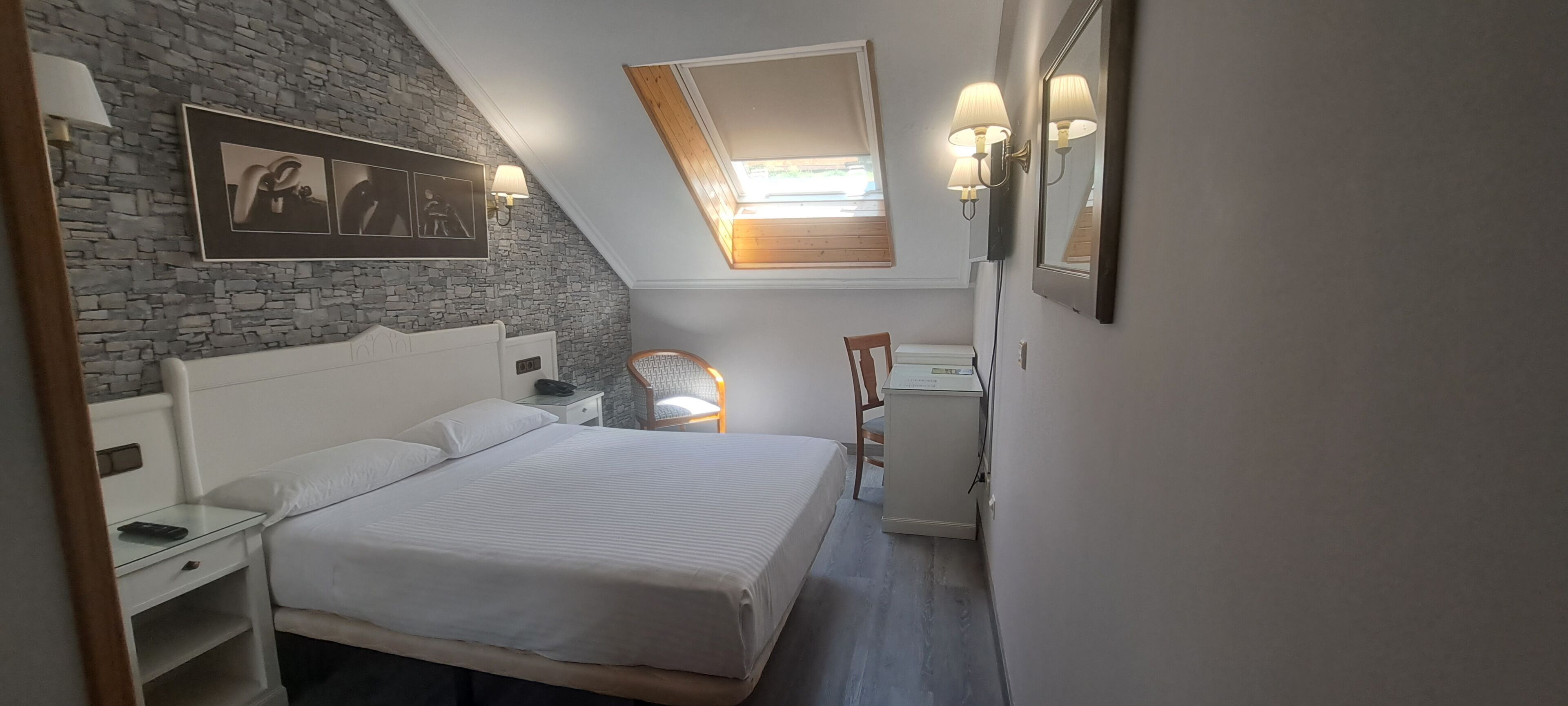 Un dormitorio con una cama grande, un escritorio, una silla, un pequeño armario y un techo inclinado con tragaluces.