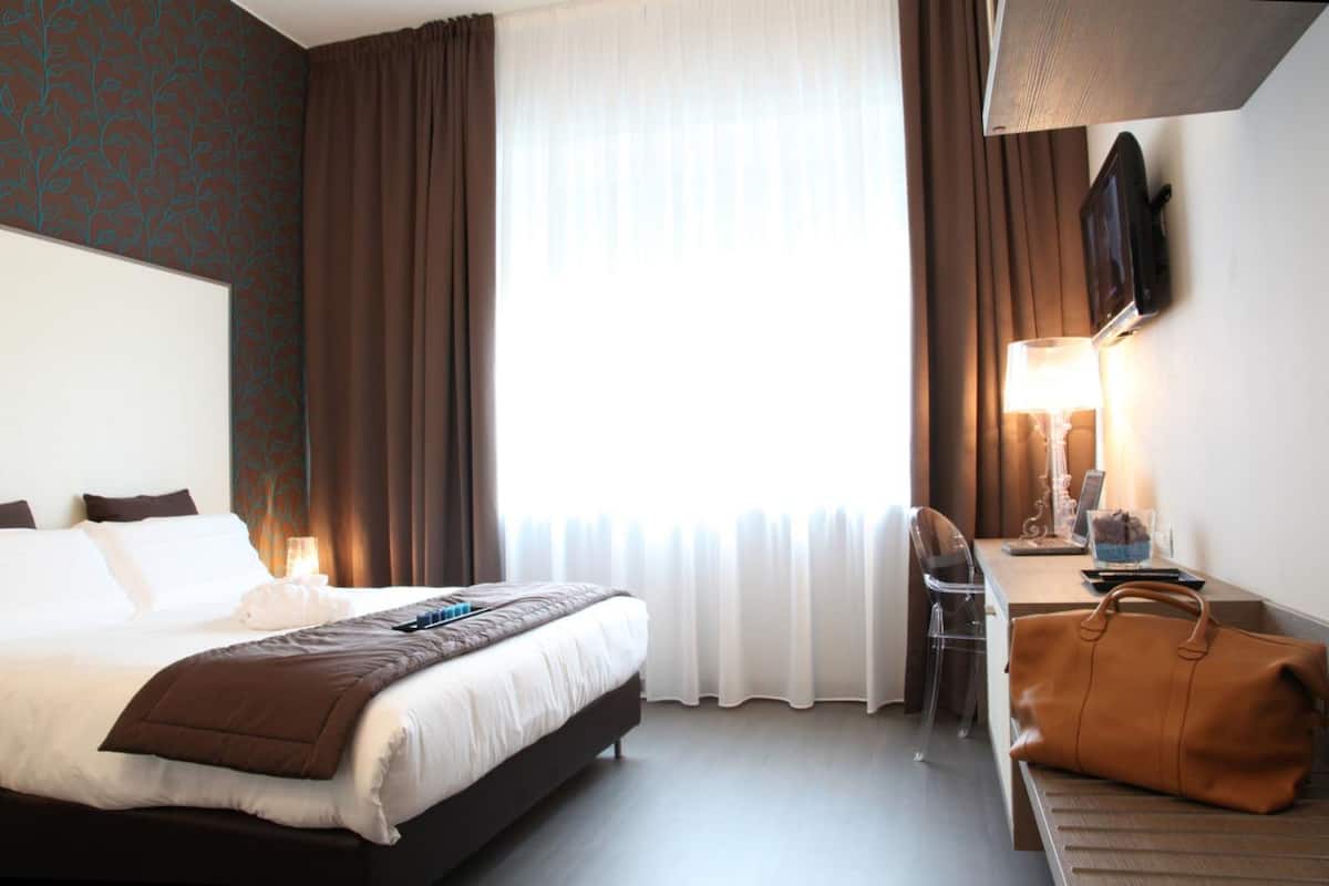 Hotellihuone, jossa on sänky, työpöytä lampun kanssa, tuoli ja suuri ikkuna, jossa on verhot.