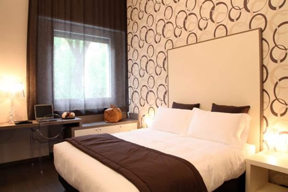 Hotellihuone, jossa on sänky, työpöytä, tuoli, kannettava tietokone ja ikkuna, jossa on verhot.
