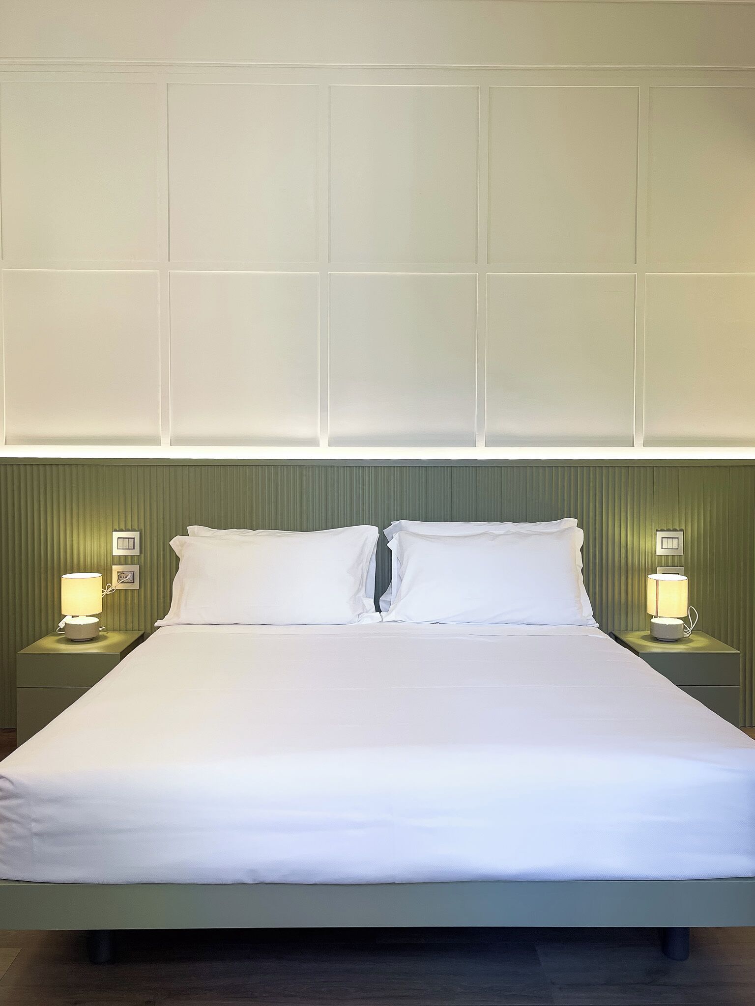 Een net opgemaakt bed met witte lakens, twee bedlampjes en een groen hoofdeinde.