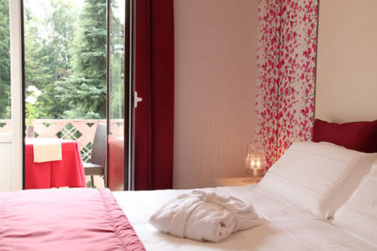 Hotellihuone, jossa on sänky, punainen verho, ikkuna, josta avautuu näkymä puihin, sekä pöytä punaisella pöytäliinalla huoneen ulkopuolella.