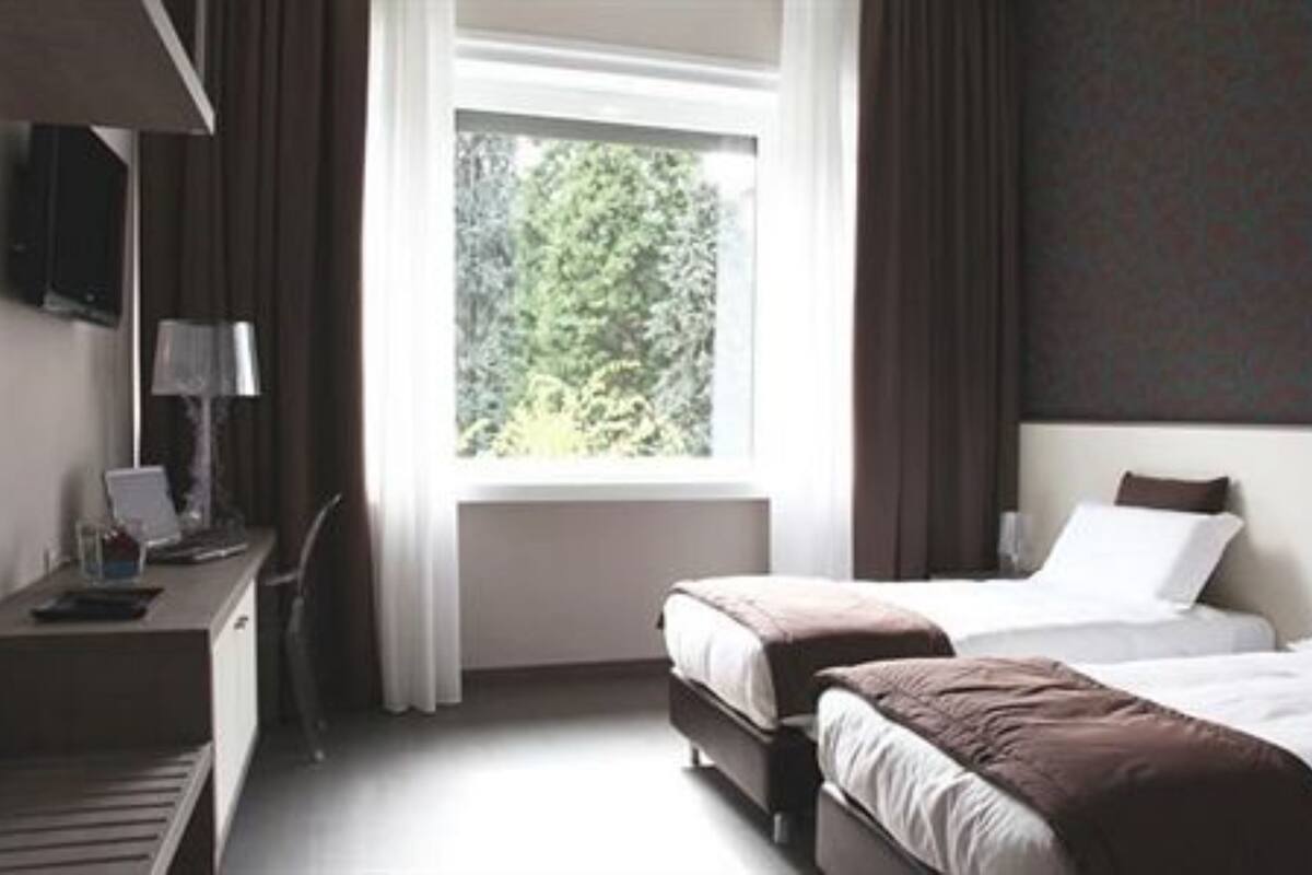 Hotellihuone, jossa on kaksi sänkyä, työpöytä lampun kanssa ja ikkuna, josta avautuu näkymä vehreään maisemaan.