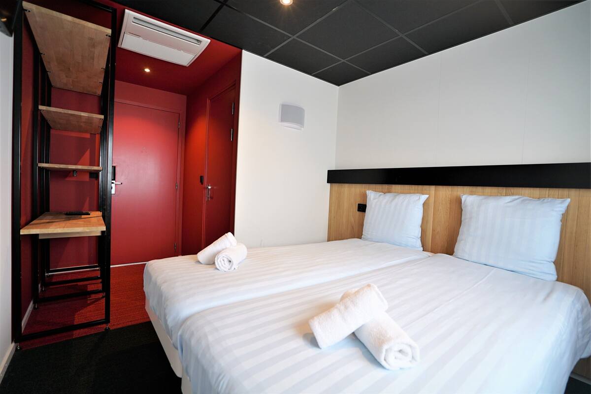 Une chambre d’hôtel avec un lit, une porte rouge, une étagère en bois et un mur blanc.