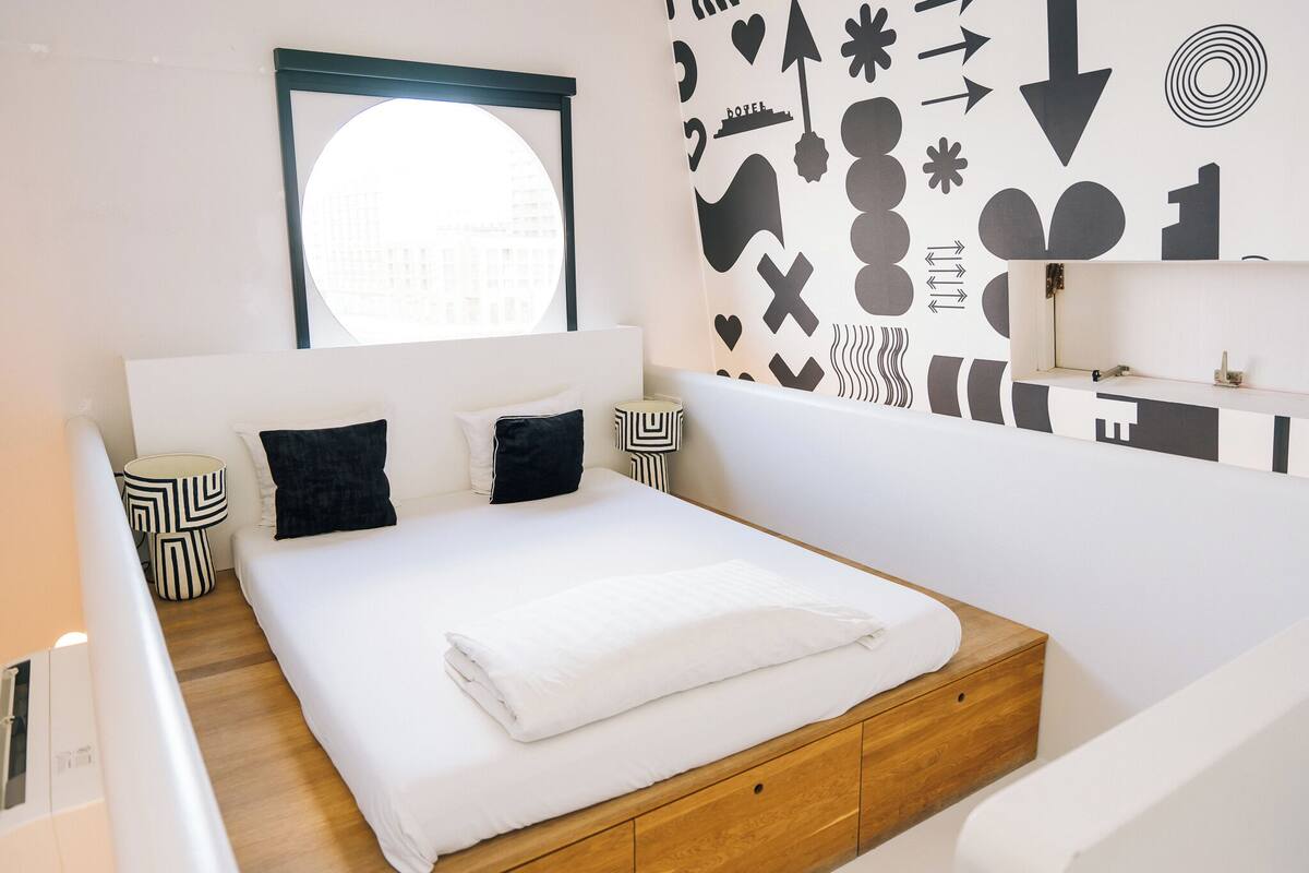Une chambre moderne avec un grand lit, une tête de lit en bois, une fenêtre ronde et un mur orné de motifs abstraits en noir et blanc.