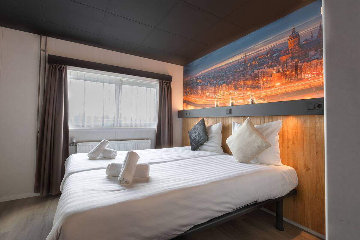 Une chambre d’hôtel avec un grand lit, un tableau représentant un paysage urbain accroché au mur, une fenêtre avec des rideaux et un radiateur.