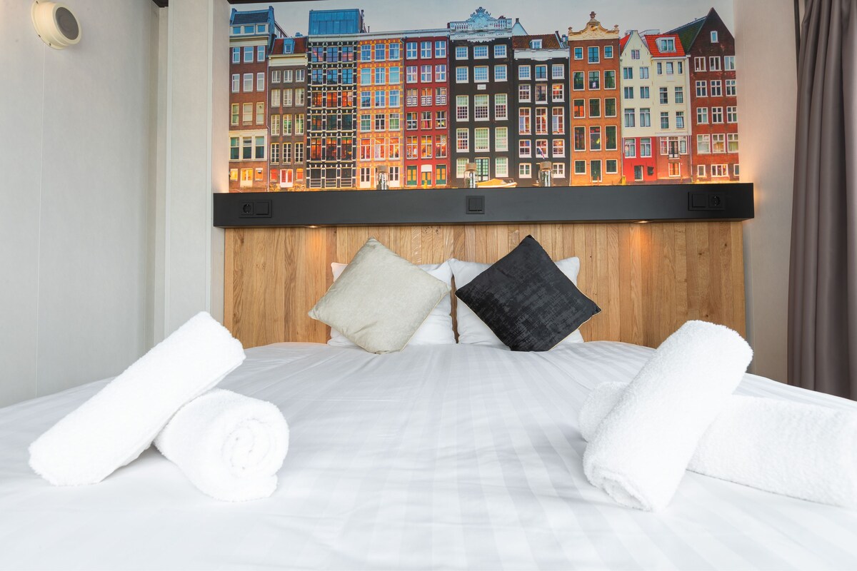 Habitación de hotel con cama, ropa de cama blanca, dos almohadas y un mural de edificios coloridos.