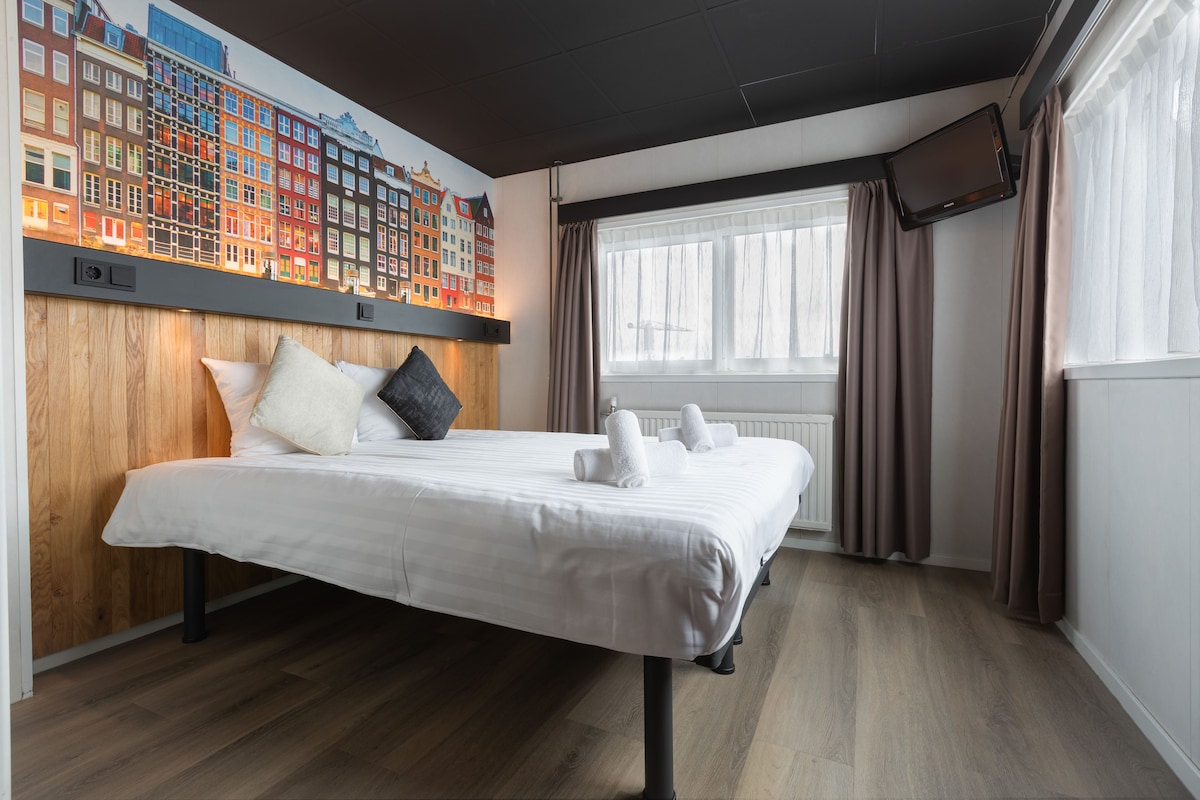 Habitació d'hotel amb un llit gran, amb capçal de fusta, un mural de paisatge urbà, una finestra amb cortines i una televisió de pantalla plana penjada a la paret.