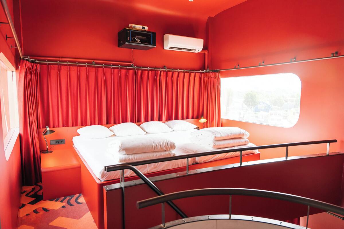 Une chambre d’hôtel de taille modeste, avec un lit simple, des rideaux rouges, une petite table de chevet avec une lampe et un appareil de climatisation fixé au mur.