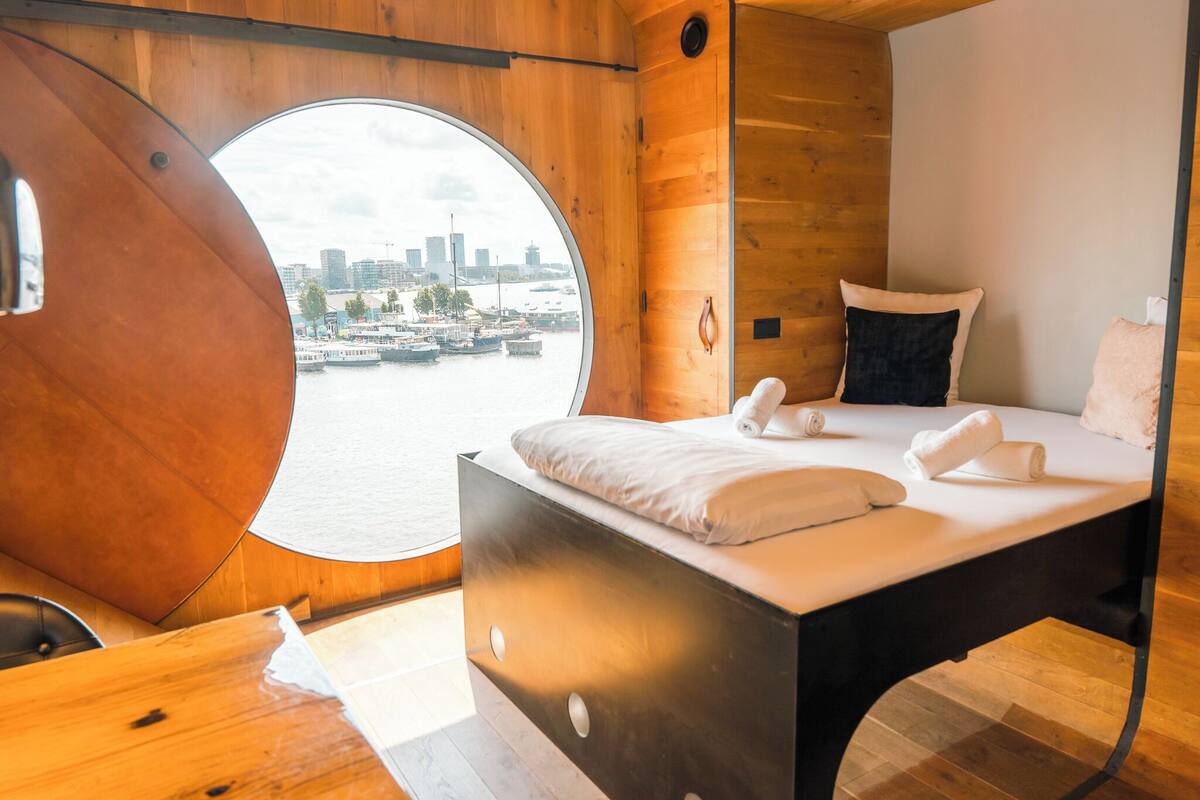 Une chambre moderne dotée d’une fenêtre circulaire, d’un lit avec du linge de lit blanc et d’un intérieur en bois.
