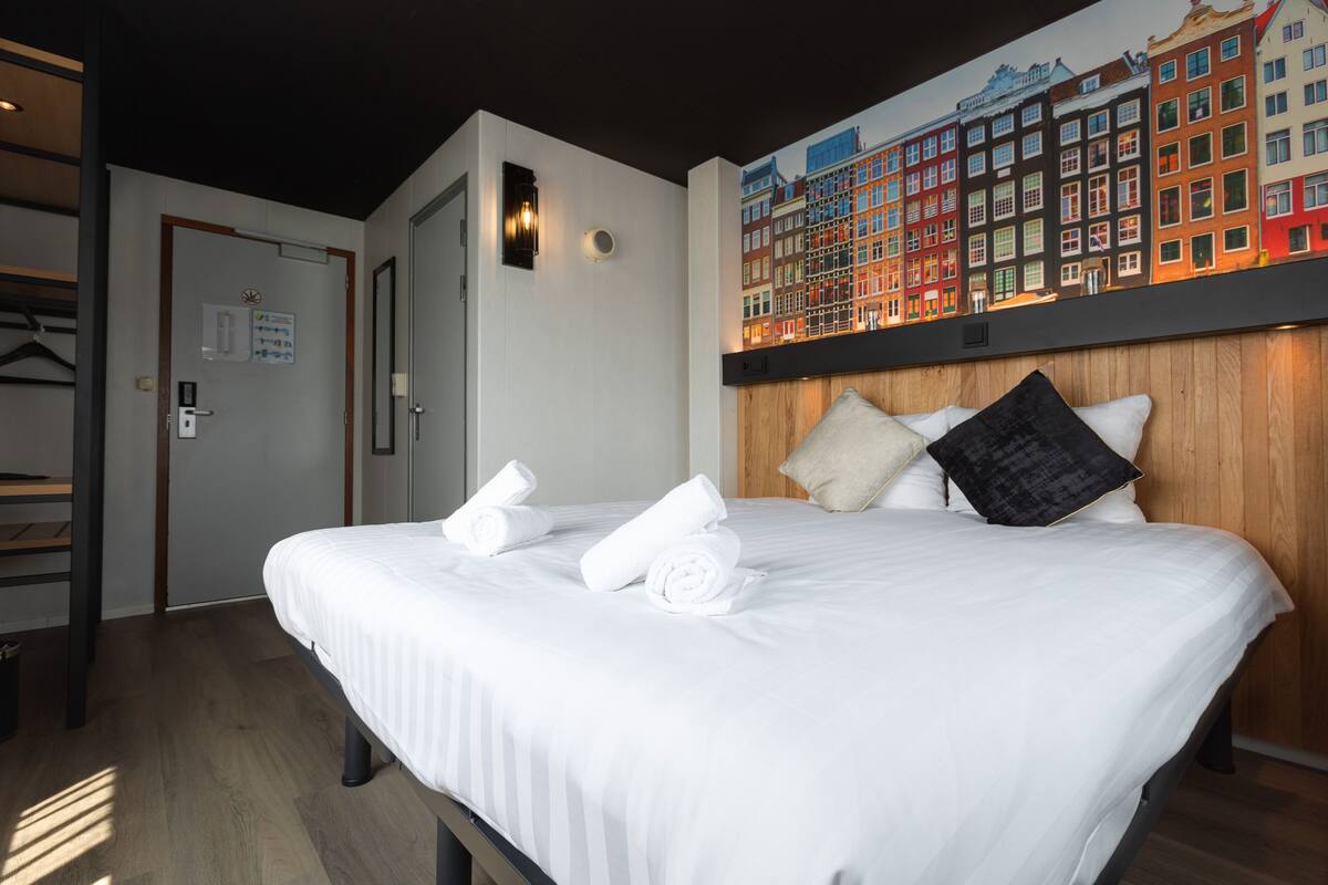 Une chambre d’hôtel avec un lit bien fait, une tête de lit en bois et une fresque murale représentant des bâtiments aux couleurs vives.