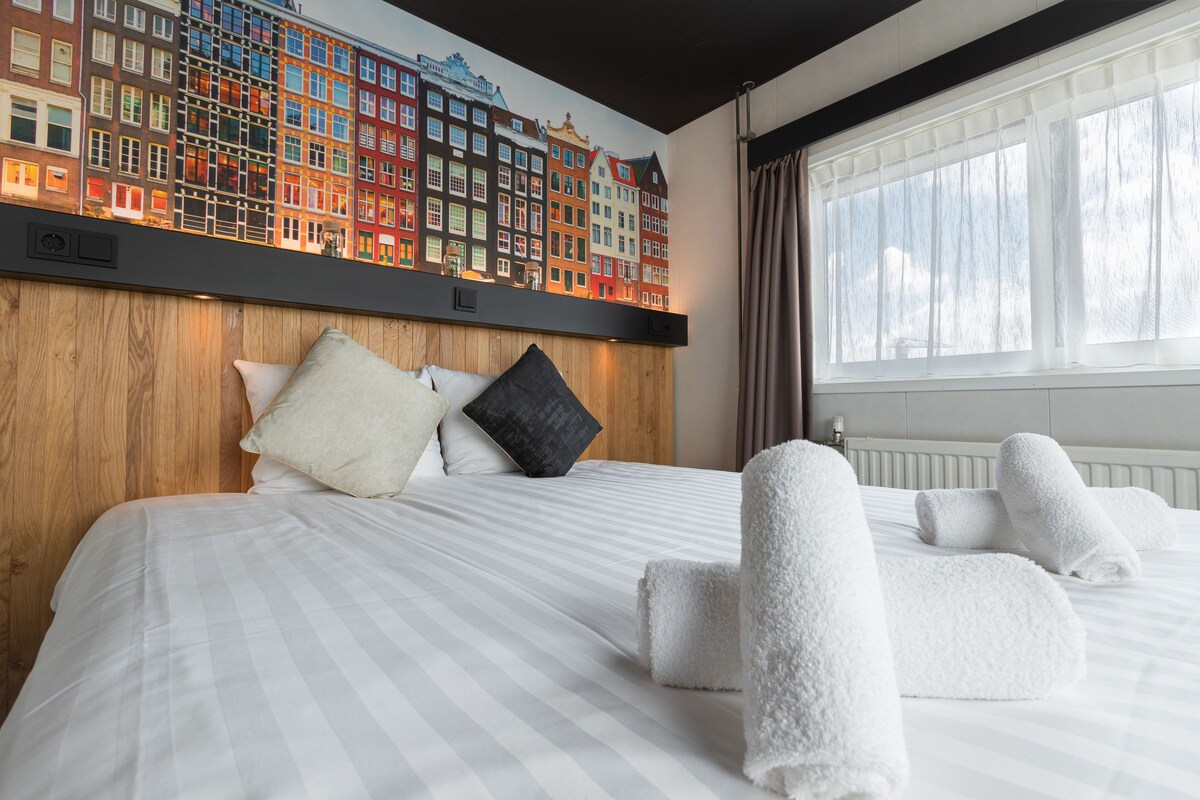 Una habitació d'hotel amb un llit gran, un panell de fusta al capçal, un mural de paisatge urbà i tovalloles ben arremegades.