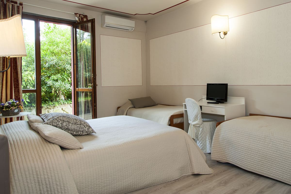 A hotel room with two beds, a desk with a TV, and a window with a view of greenery.