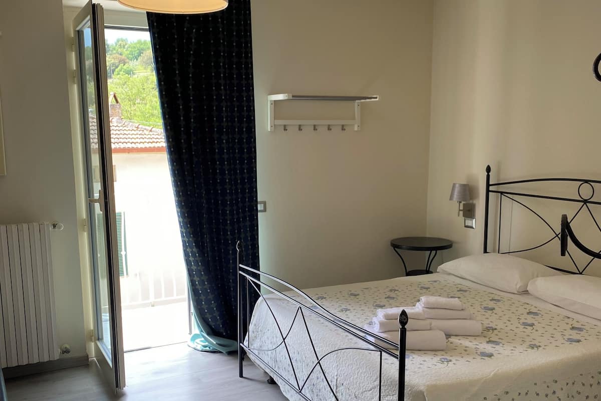 A bedroom with a bed, a bedside table, a hanging light fixture, a window with curtains, and a view of the outside.