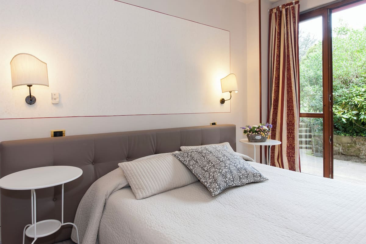 A bedroom with a bed, two wall-mounted lamps, a small round table, and a view of greenery through a window.