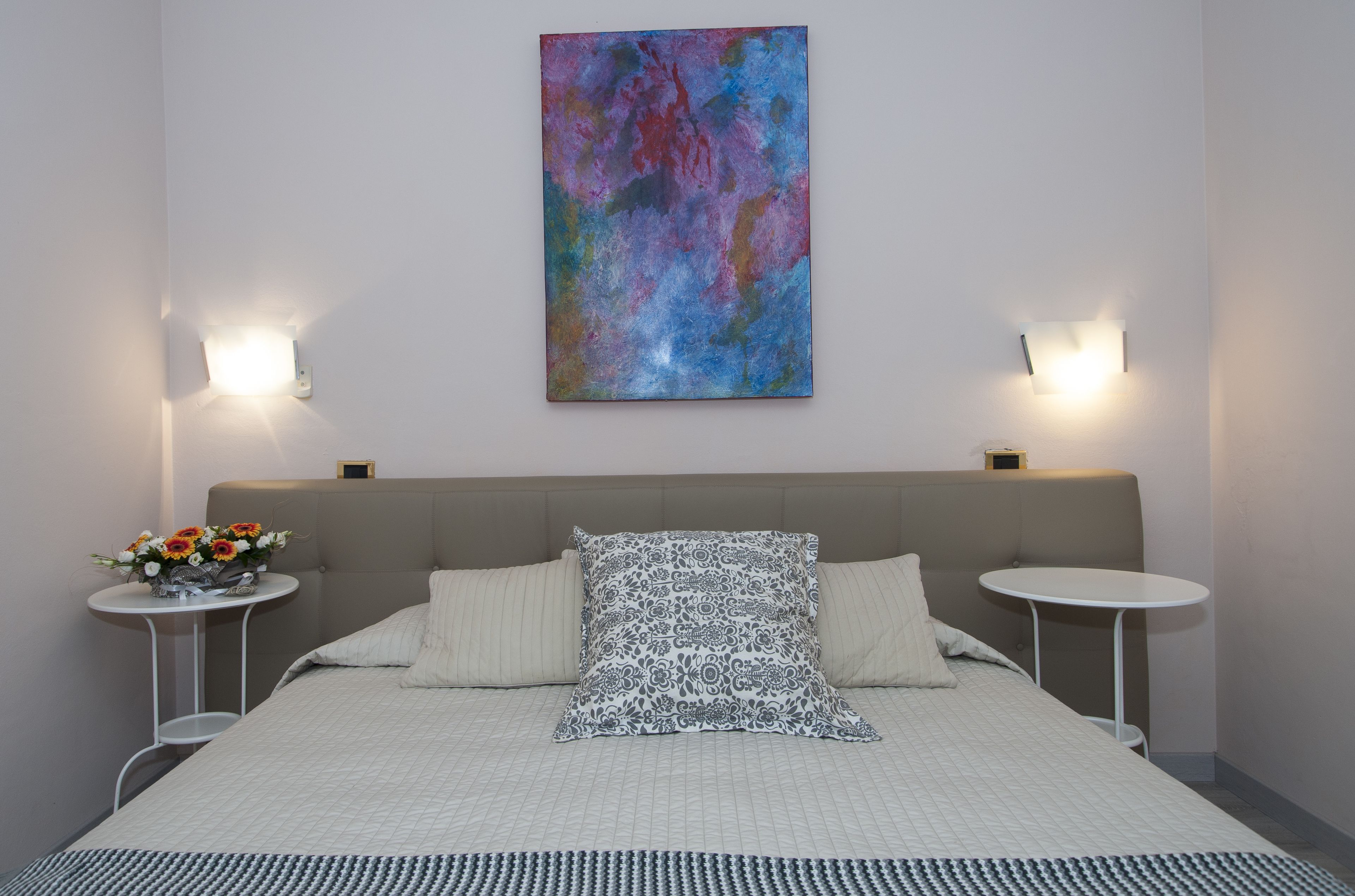 Une chambre d’hôtel moderne avec un grand lit, deux tables de chevet avec des lampes, un élément décoratif mural et une petite table ronde agrémentée de fleurs.