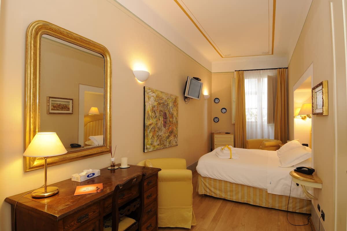 Habitación de hotel con un espejo grande, un escritorio con lámpara, una cama y un televisor.