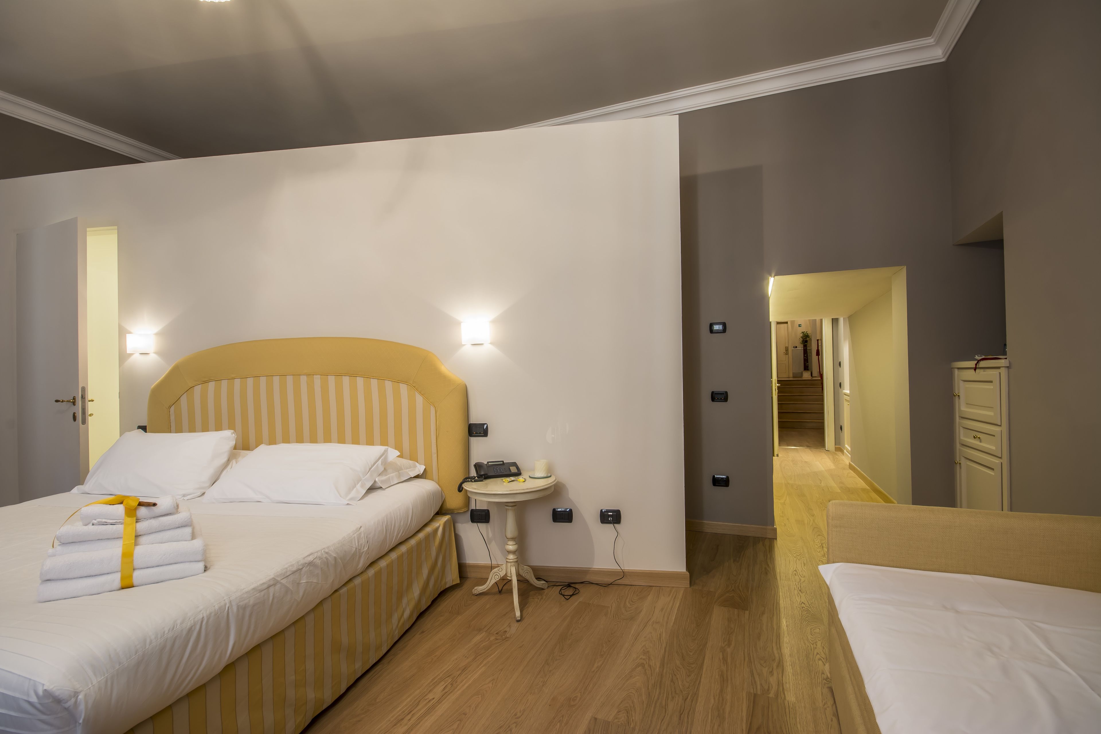 Habitación de hotel con una cama, mesita de noche y una pequeña mesa con lámpara. Se observa una puerta que conduce a otra habitación y un pasillo con una escalera.