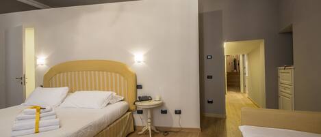 Habitación de hotel con una cama, mesita de noche y una pequeña mesa con lámpara. Se observa una puerta que conduce a otra habitación y un pasillo con una escalera.