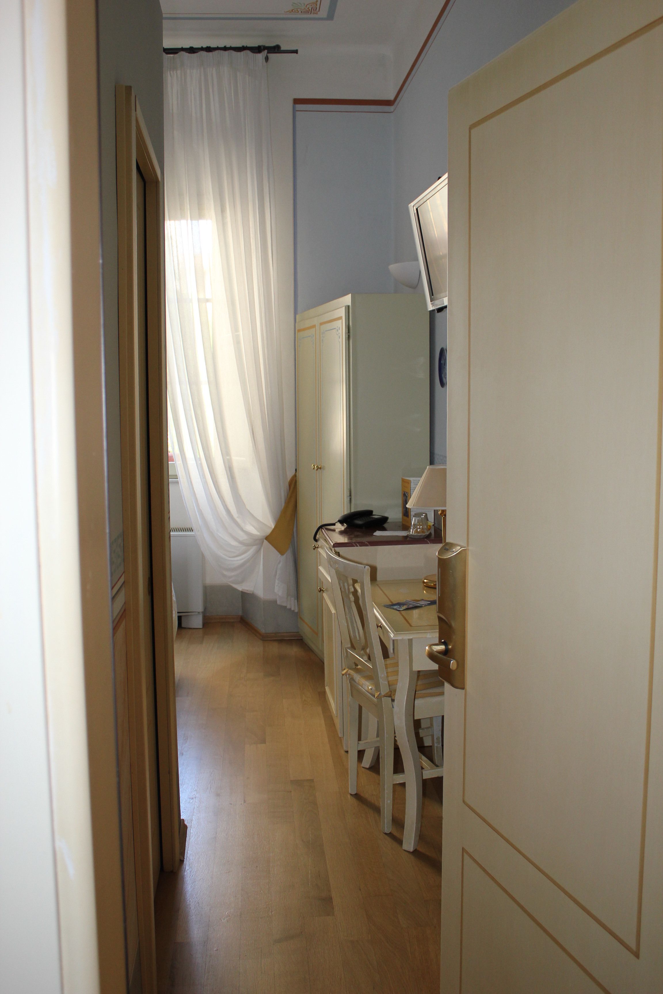 A narrow hallway with a wooden floor, a white door on the right, and a window with sheer curtains.