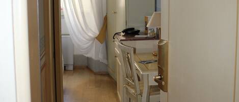 A narrow hallway with a wooden floor, a white door on the right, and a window with sheer curtains.