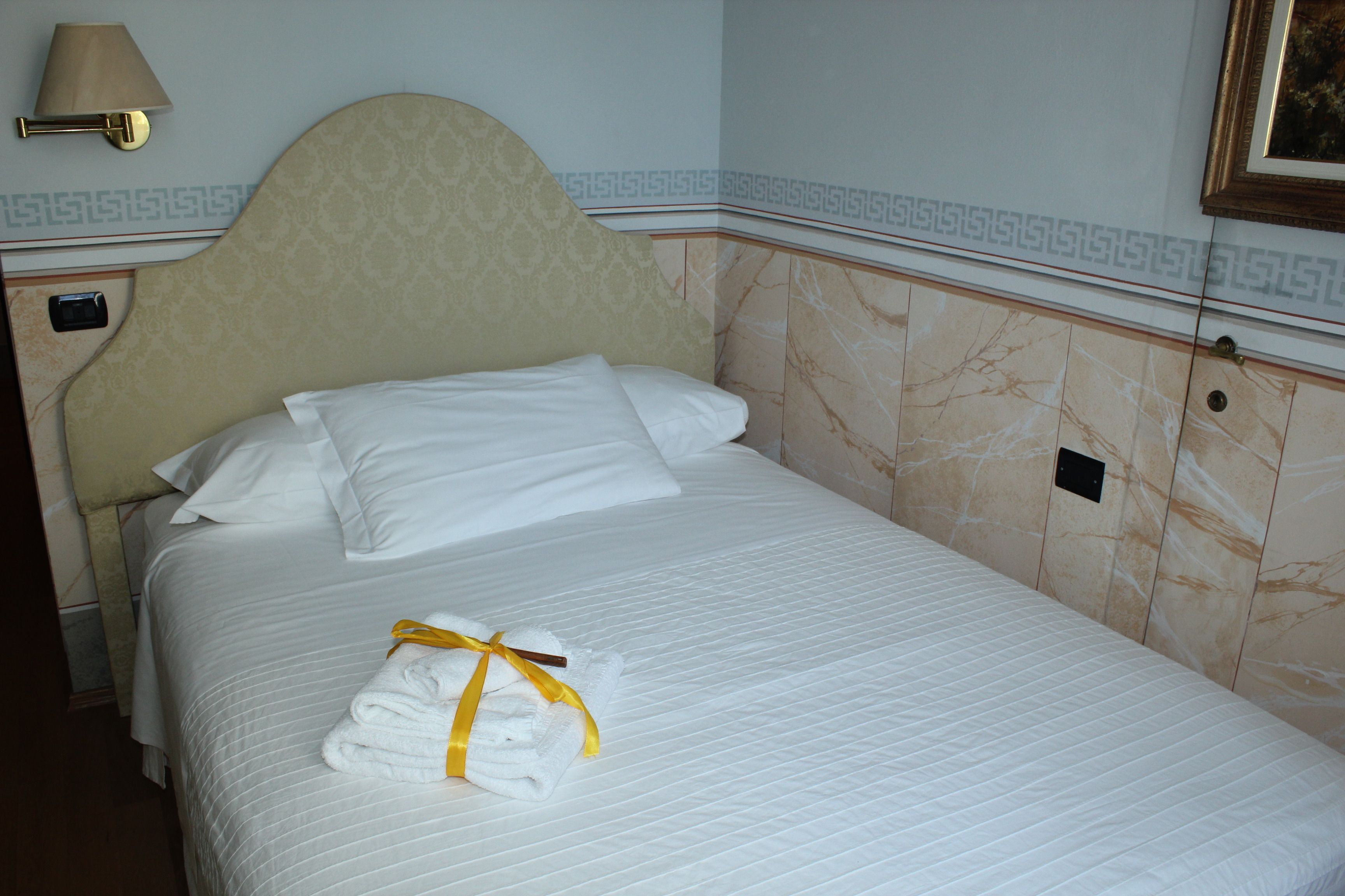 A neatly made bed with white linens and a yellow towel, a headboard with a decorative pattern, and a wall-mounted lamp.