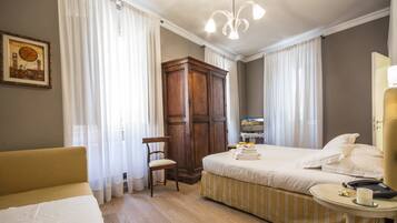 A hotel room with a bed, a chair, a wardrobe, a bedside table with a phone, and a painting on the wall.