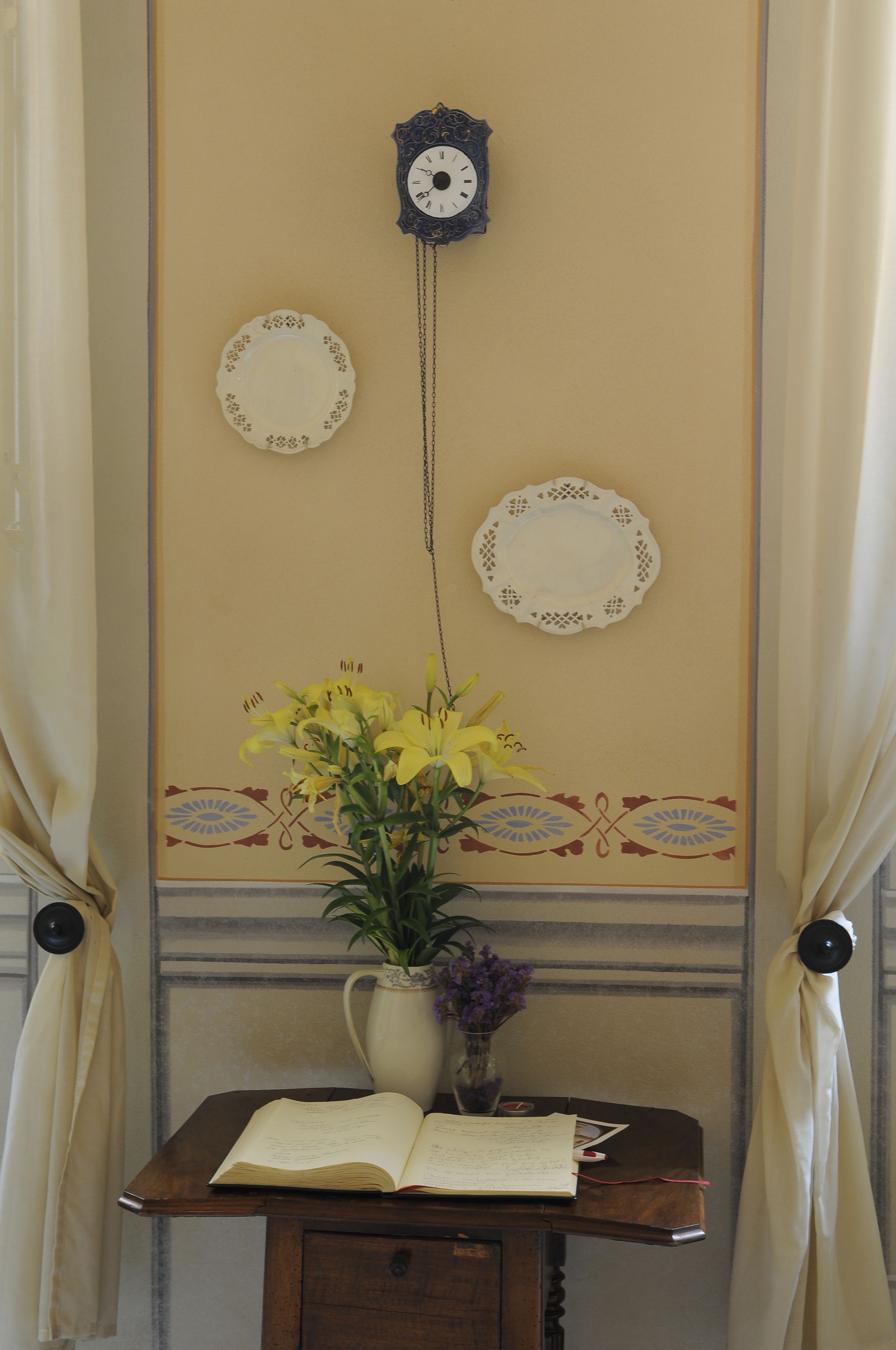 A room with a clock on the wall, a vase with flowers, and a table with an open book.
