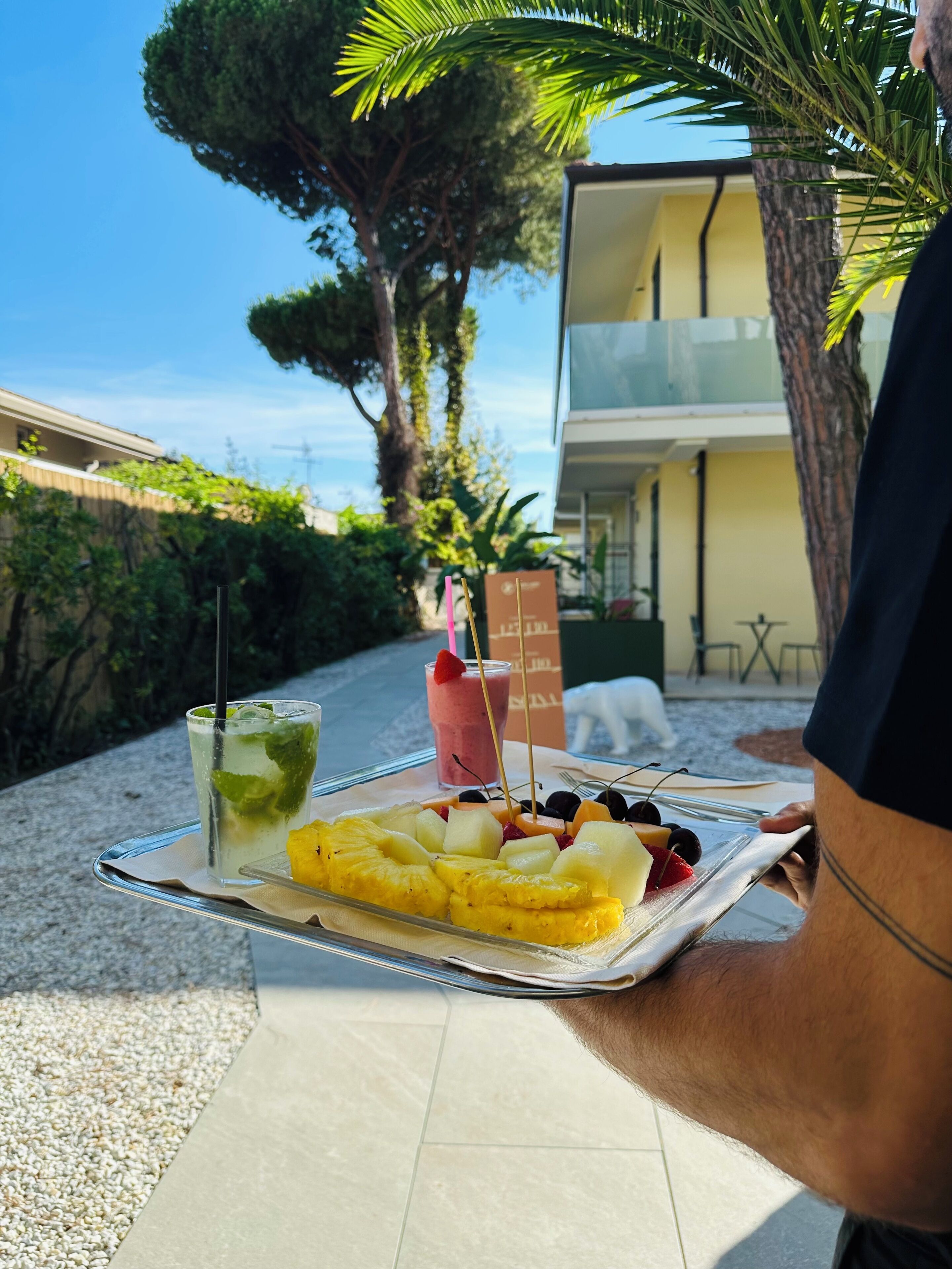 A tray with a fruit platter and drinks on a terrace with a view of a building and trees.