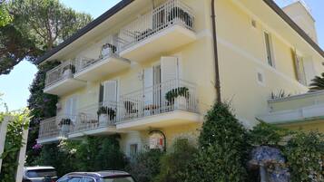 Un edificio a più piani con balconi, un'auto parcheggiata di fronte e vegetazione rigogliosa.